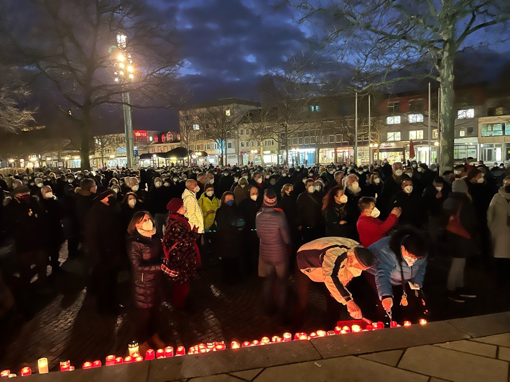 Das Foto zeigt eine Veranstaltung vor dem Wolfsburger Rathaus. Auf der Treppe stehen Grabkerzen, von denen jede einzelne für einen Corona-Toten dieser Stadt steht. Zwei Menschen zünden die Kerzen an, im Hintergrund sind viele Bürger zu sehen. 